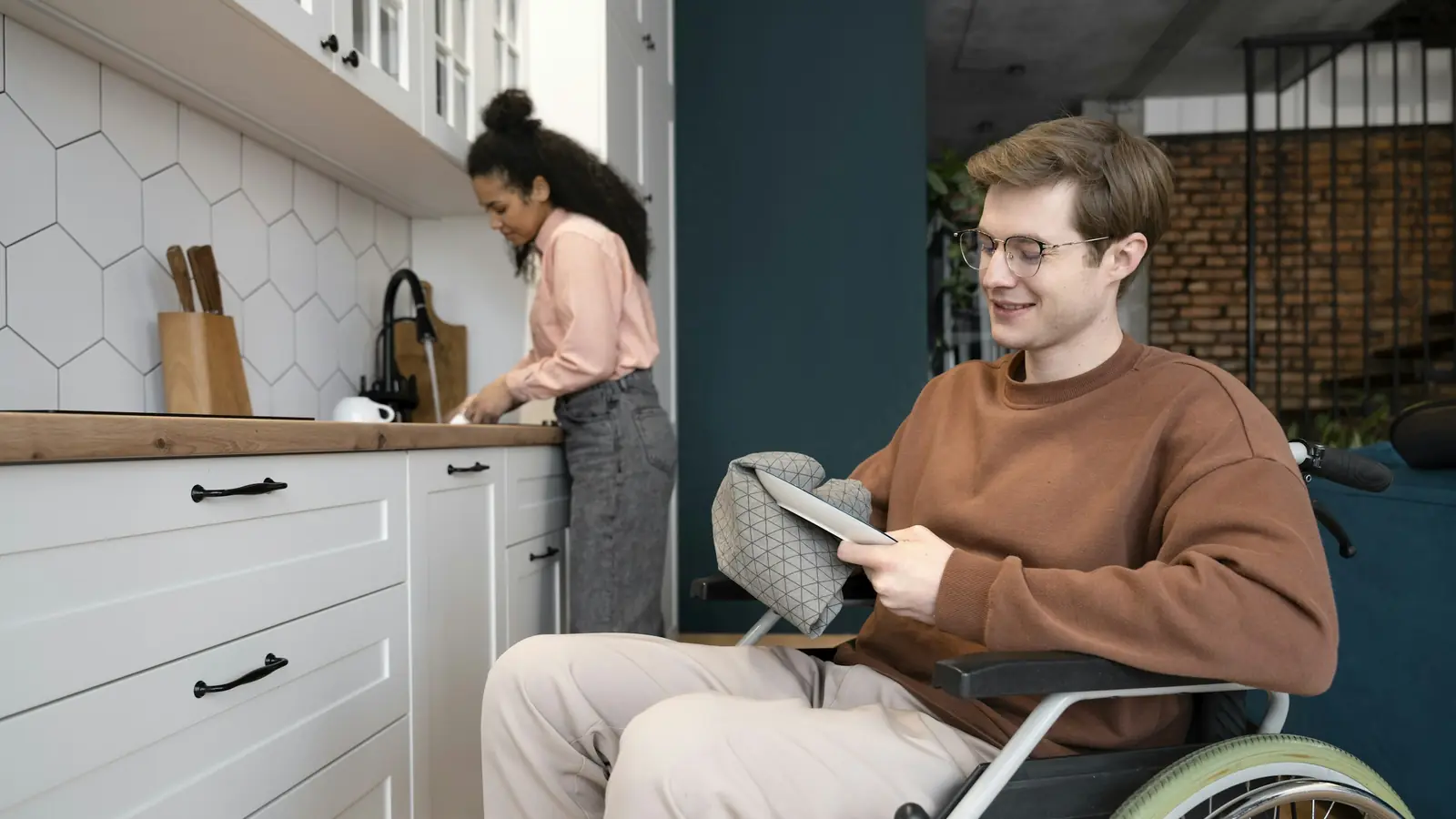 Ein junger Mann im Rollstuhl schaut in seiner Küche auf ein Tablet, im Hintergrund steht eine Frau an der Spüle.