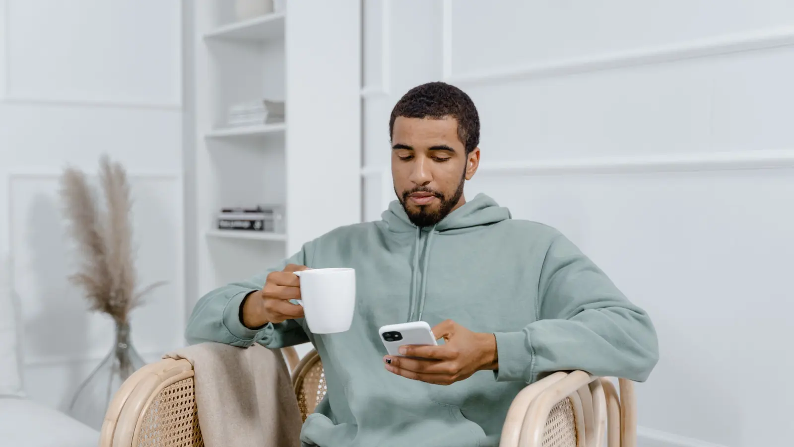 Ein junger Mann im Kapuzenpullover sitzt mit einer Tasse in einem Sessel und liest eine Nachricht auf seinem Smartphone.