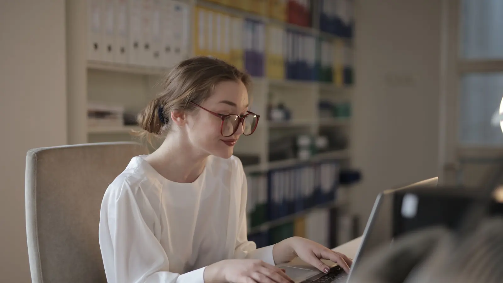 Eine junge Frau mit Brille arbeitet in einem Büro mit Aktenregalen am Laptop.