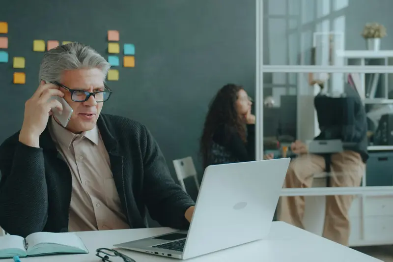 Ein Mann mit Brille telefoniert am Schreibtisch und schaut dabei auf seinen Laptop.