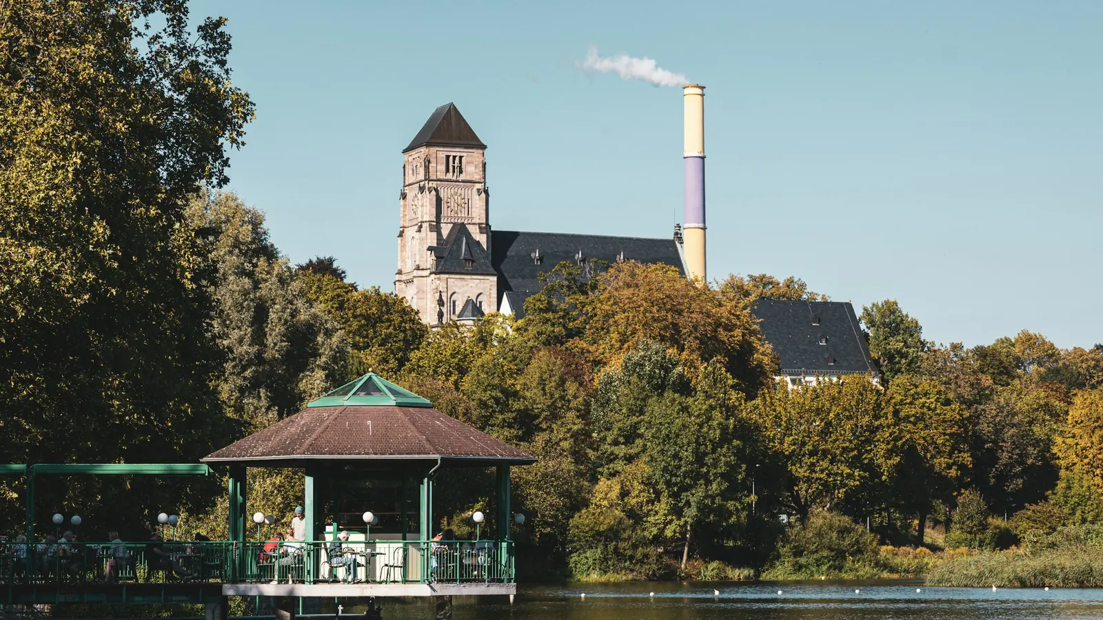 Der Schloßteich in Chemnitz im Herbst mit einem Pavillon am Wasser, dahinter der Turm der Schlosskirche und die gelb und violett gestreifte Esse.