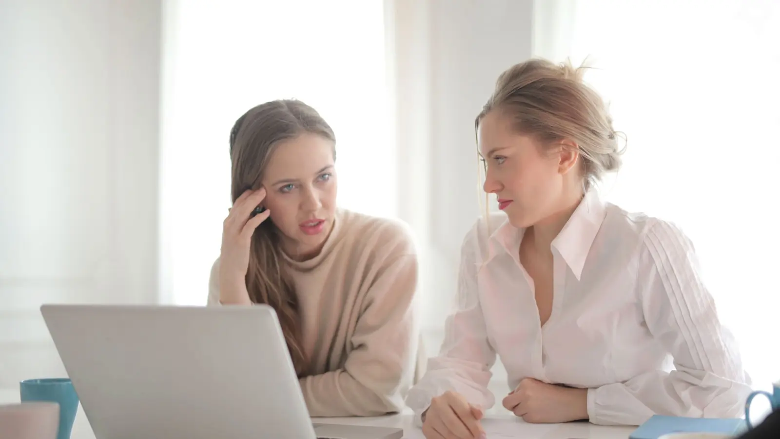 Zwei Frauen sitzen an einem Tisch mit Laptop und besprechen nachdenklich, was sie gerade gelesen haben.