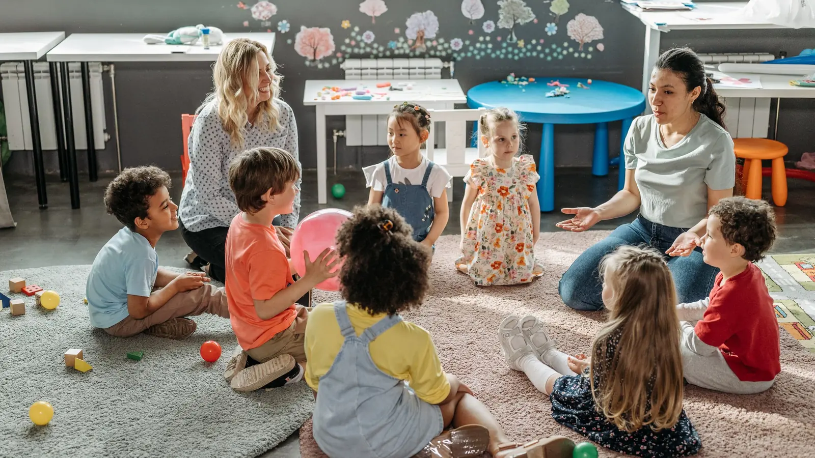 Kinder sitzen mit zwei Erzieherinnen im Kreis auf dem Teppich einer Kita.
