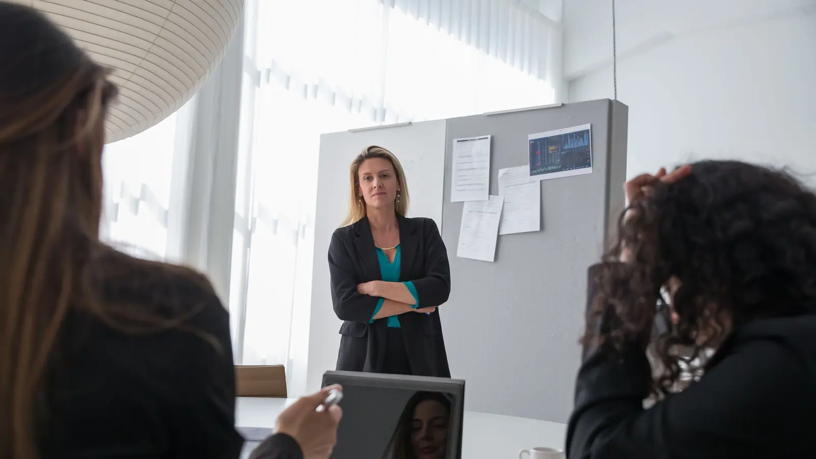 Eine Frau steht in einem Besprechungsraum vor einer Stellwand, zwei Personen sitzen im Vordergrund am Tisch und hören zu.
