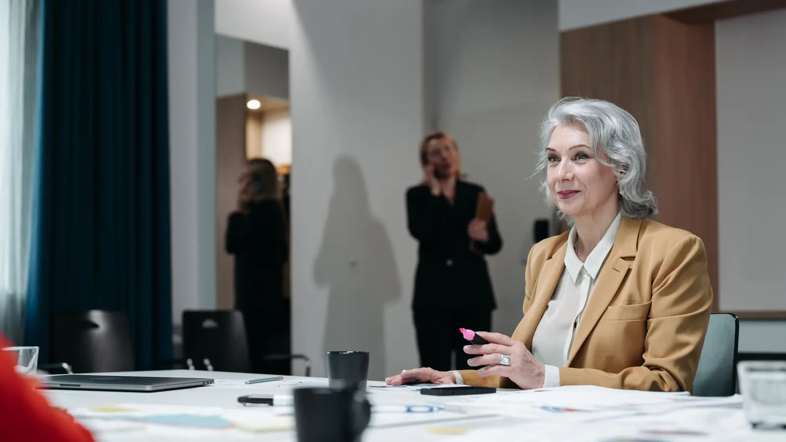 Eine Frau mit grauem Haar und senfgelbem Blazer sitzt mit Unterlagen am Besprechungstisch, im Hintergrund telefoniert eine Kollegin.
