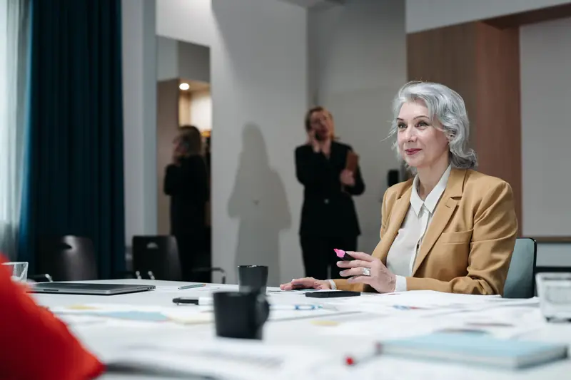 Eine Frau mit grauem Haar und senfgelbem Blazer sitzt mit Unterlagen am Besprechungstisch, im Hintergrund telefoniert eine Kollegin.
