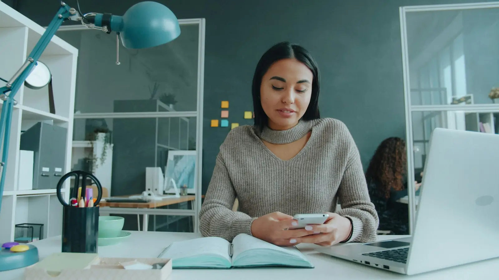 Eine Frau sitzt im Büro am Schreibtisch und tippt auf ihrem Smartphone, daneben ein Laptop.