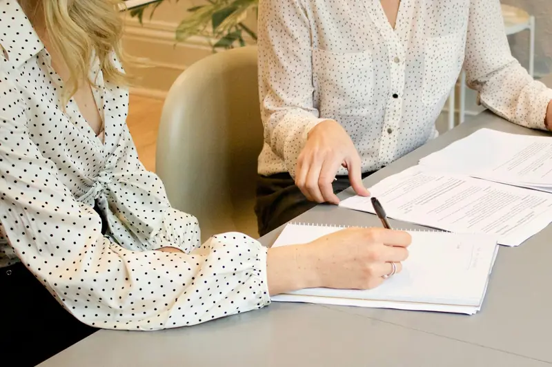 Zwei Frauen gehen an einem Tisch Vertragsunterlagen durch, eine macht sich Notizen.