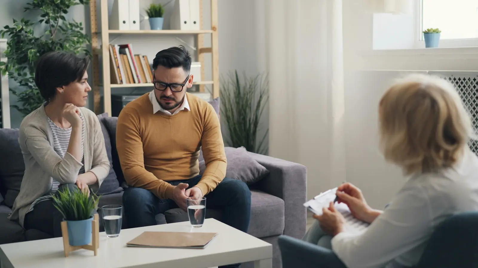 Ein Paar sitzt auf dem Sofa eines Beratungsraums, eine Beraterin mit Schreibbrett hört zu.