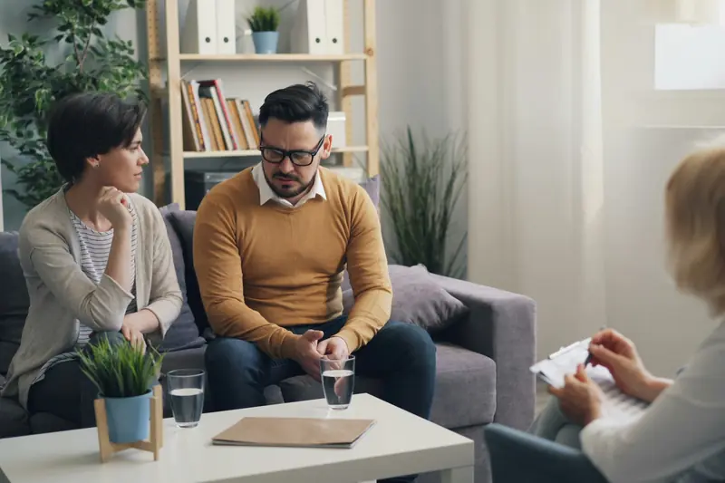 Ein Paar sitzt auf dem Sofa eines Beratungsraums, eine Beraterin mit Schreibbrett hört zu.