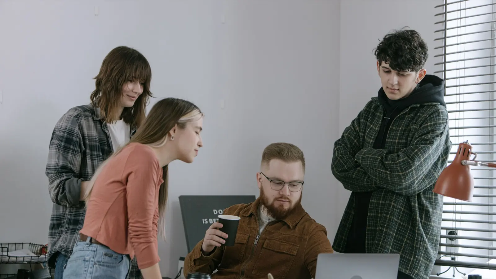 Vier junge Menschen in Alltagskleidung schauen in einem schlichten Büro gemeinsam auf einen Laptop, ein Mann sitzt in der Mitte mit einem Becher in der Hand.