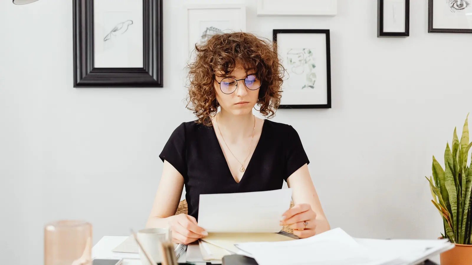 Eine Frau mit Brille sitzt an einem hellen Schreibtisch und liest ein Blatt aus einem Stapel Unterlagen.