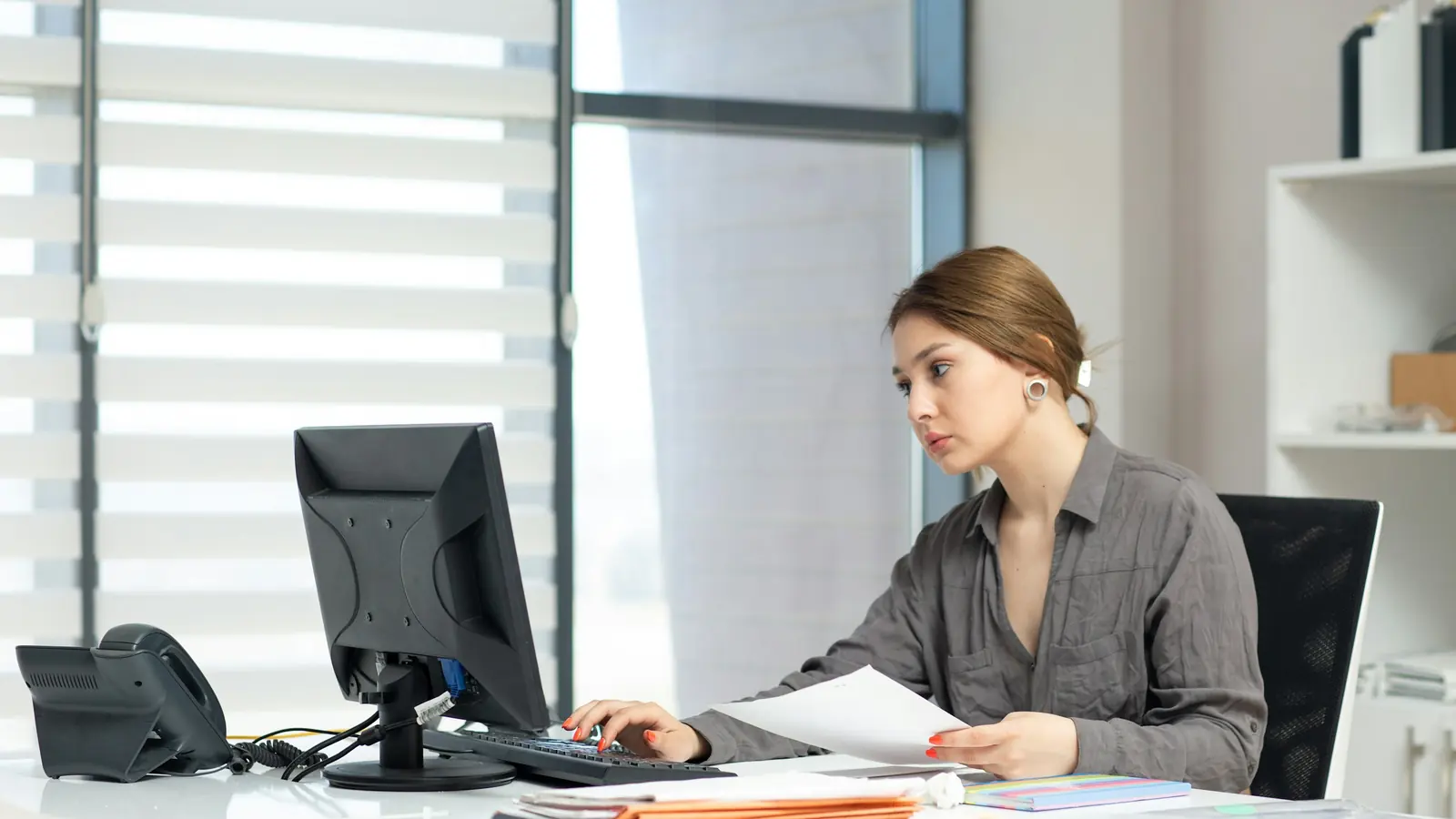 Eine Mitarbeiterin sitzt in einem hellen Büro am Computer, hält ein Blatt Papier in der Hand und tippt dabei, auf dem Schreibtisch liegen Aktenmappen, ein Telefon und ein Taschenrechner.